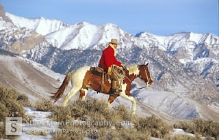 A man in a red coat rides a brown and white horse through the mountains. The sky is clear of clouds and snow sits on the peaks of the mountains in the  background