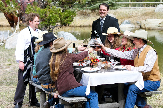 A group of guests in cowboy hats raise their wine glasses around a picnic table outside