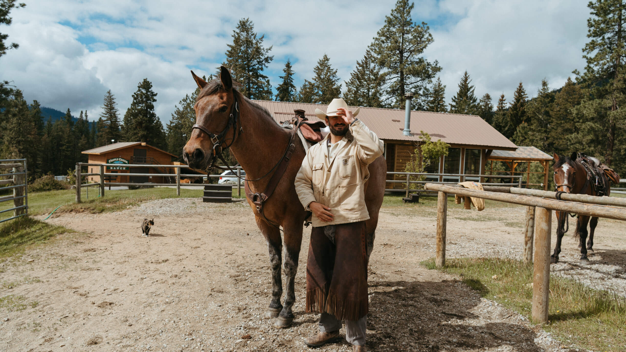 Male Guest and horse with barn cat Bonnie in the background in the arena in front of the Rider's Roost at the barn about to depart on horseback trail ride