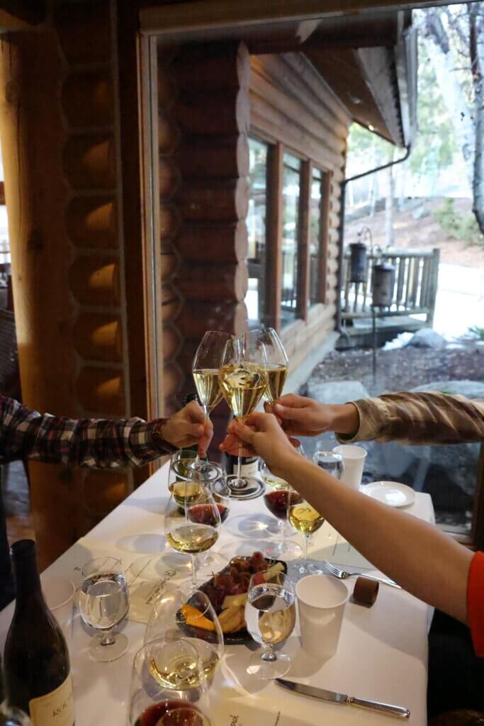 Three people clink their wine glasses together over a luxuriously table containing multiple glasses of wine and plates of fruit and cheese