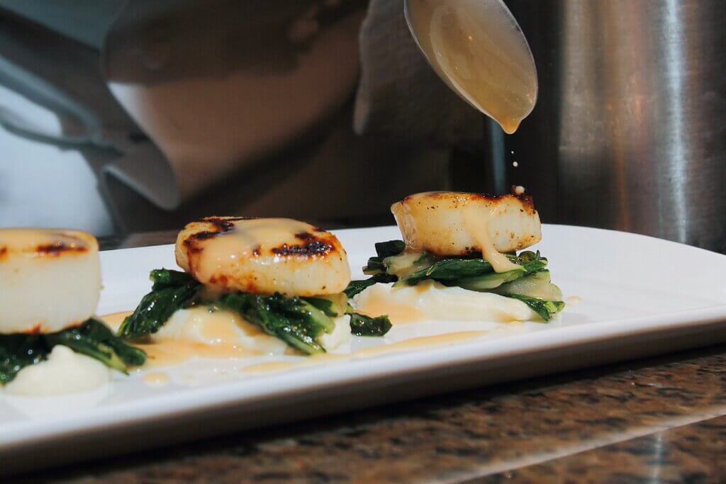 three golden-brown seared scallops rest on sautéed greens and creamy puree, with sauce being delicately drizzled from a spoon above.