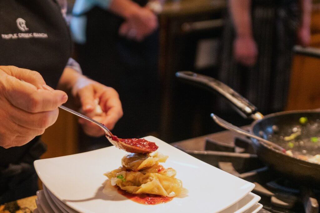 A chef in a Triple Creek Ranch apron spoons rich red sauce over a stack of dumplings or pasta on a square white plate in a warm kitchen.