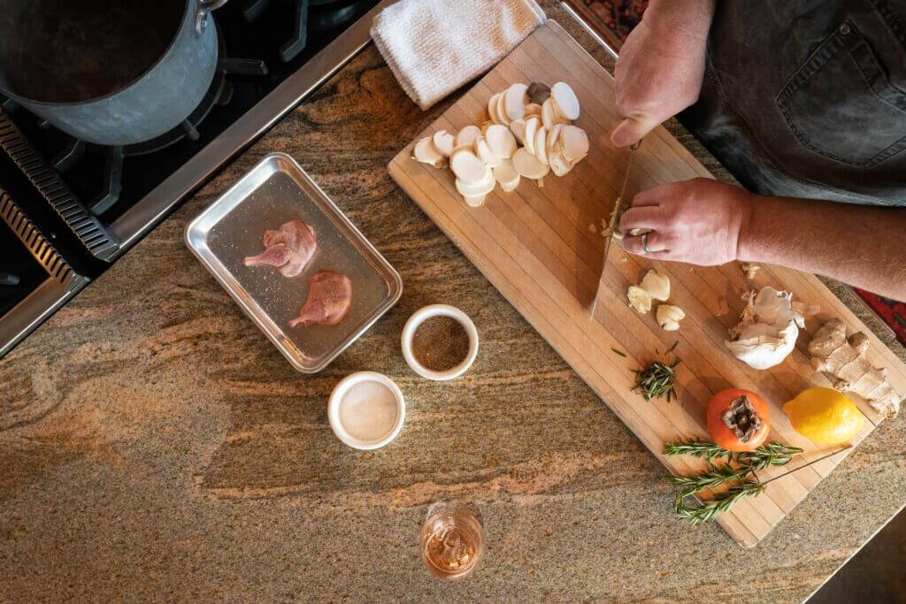Overhead view of a person chopping garlic beside sliced mushrooms, herbs, garlic, a persimmon, lemon, spices, and two raw lamb chops.