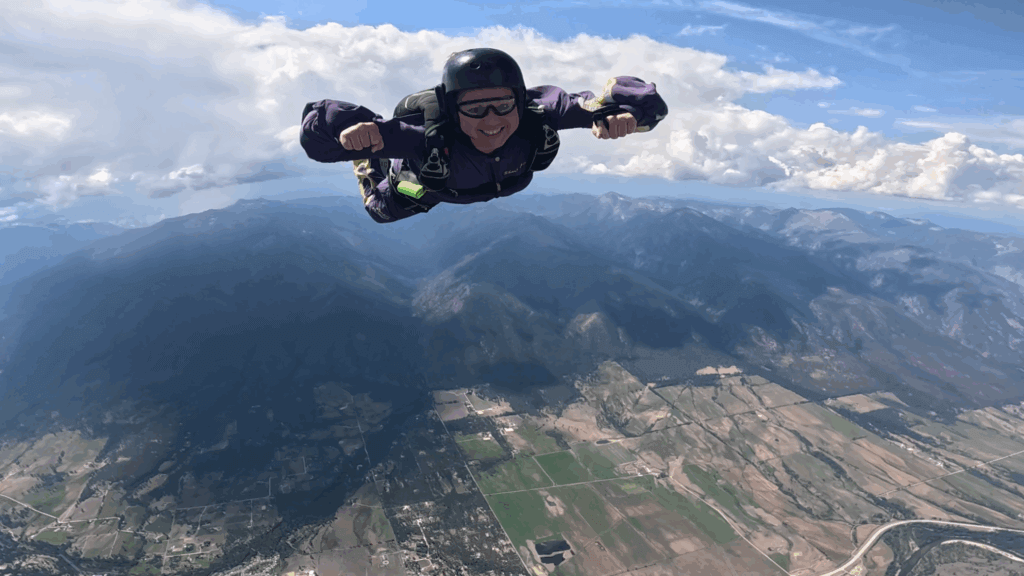 A woman in a purple skydiving suit smiles as she goes for a solo dive on a sunny day with mountains in the background