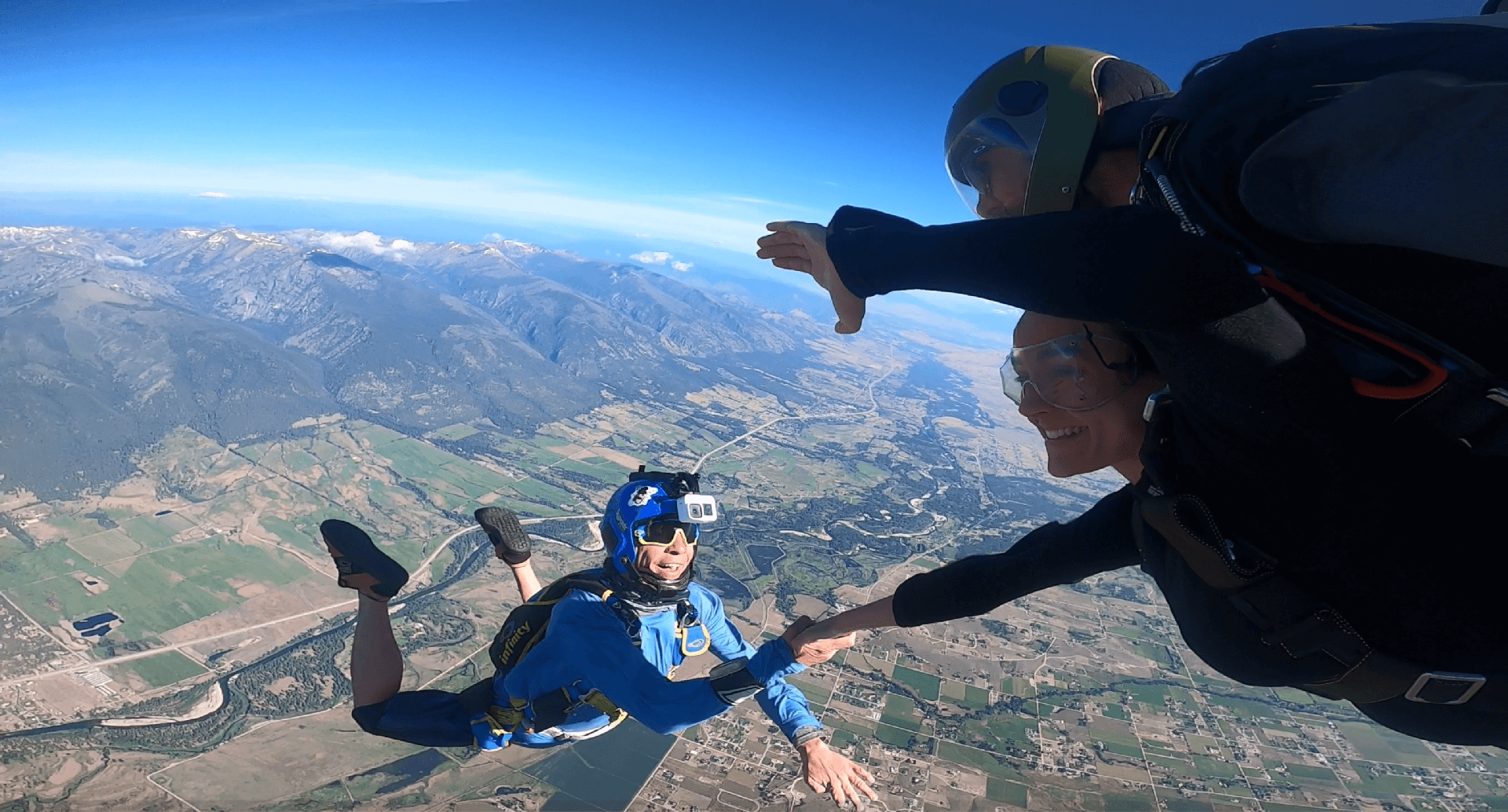 A man in a blue skydiving suit wearing a helmet mounted GoPro takes photos of a group of tandem skydivers.