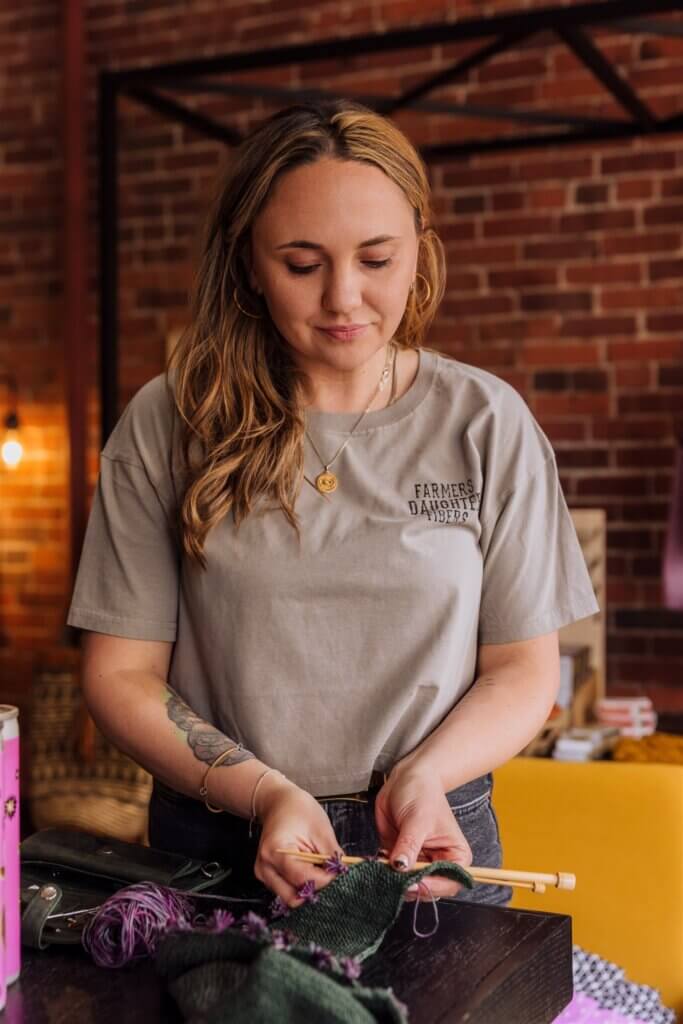 A woman in a gray t-shirt knits a green and purple patch of fabric